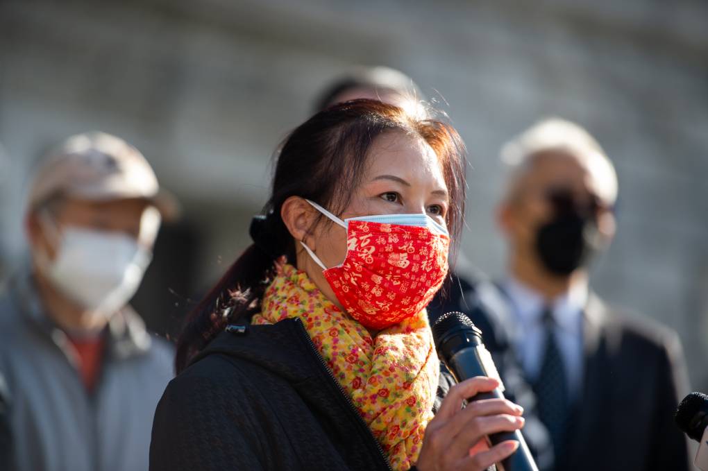 A woman wearing a red mask and yellow scarf holds a microphone.