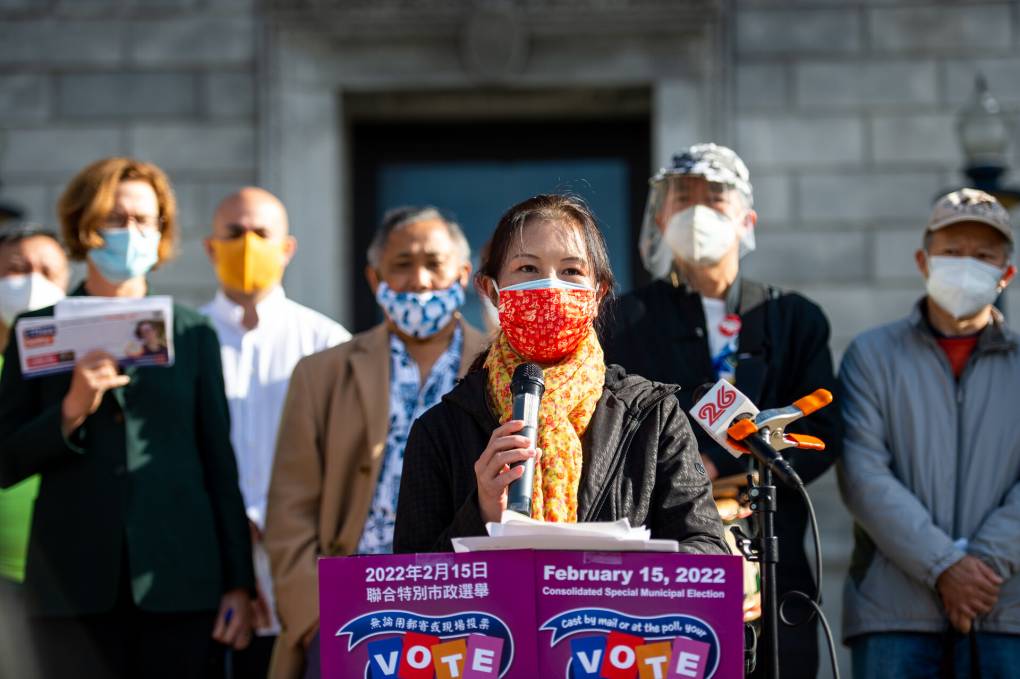 A group of seven people stand outside a building and podium wearing masks. A woman in the middle stands at the podium holding a microphone.