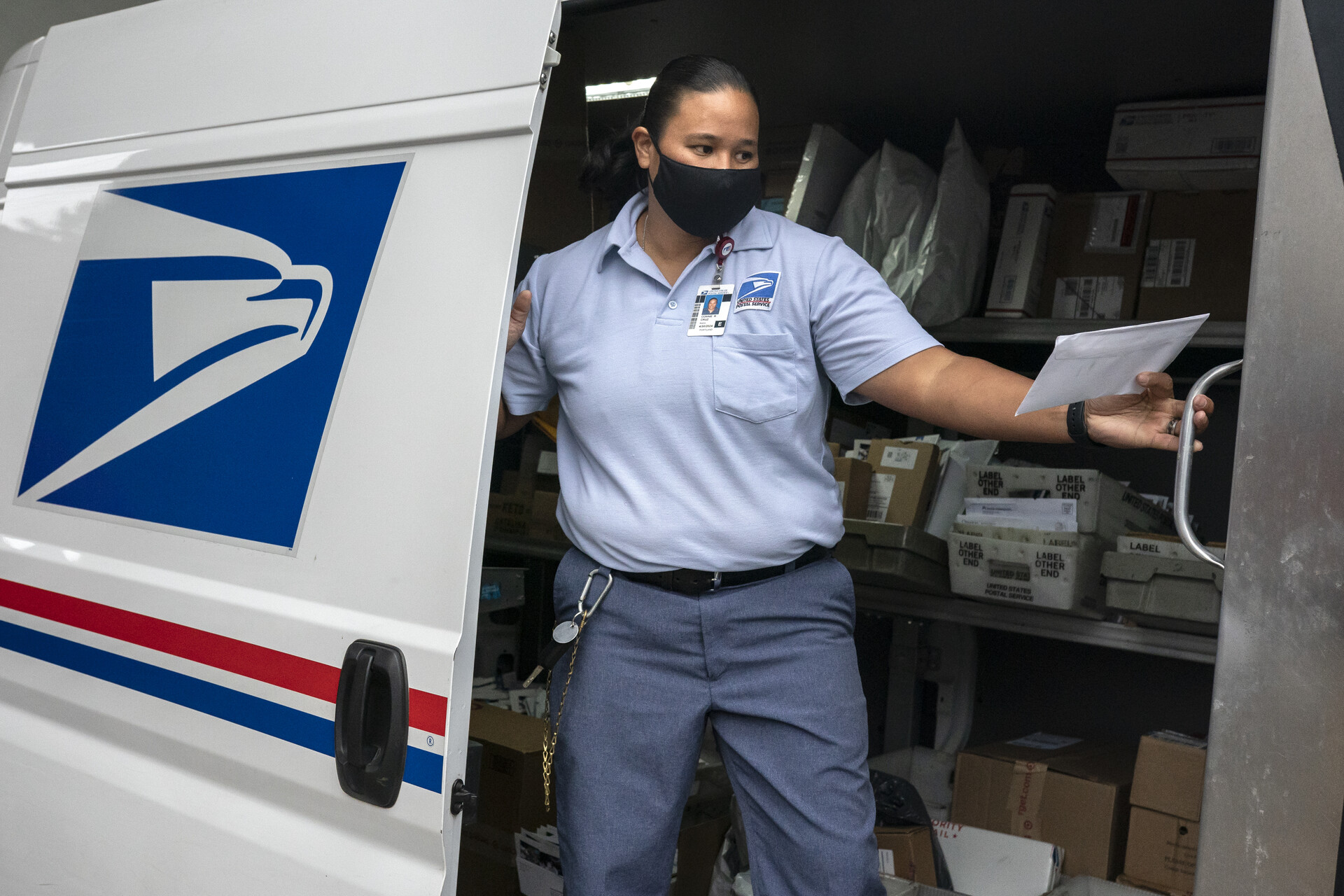 A uniformed postal service worker stands inside her mail van, with USPS logo visible on side of van