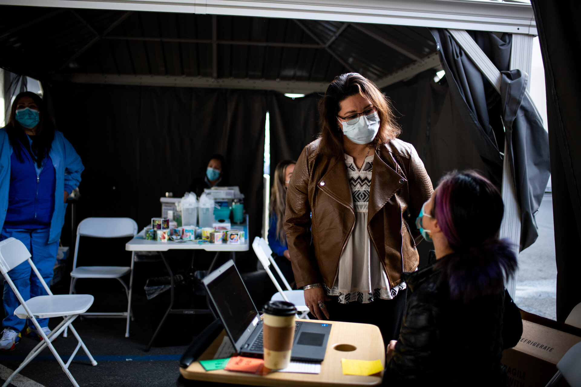 masked woman stands looking down at another masked nurse seated at a portable table, under tents at an outdoor COVID vaccination clinic