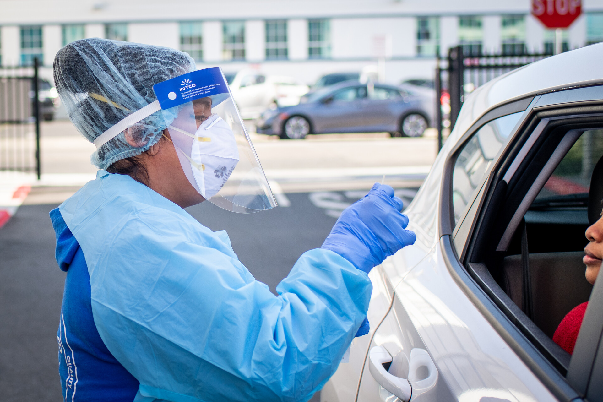nurse clothed in full PPE administers COVID test through a car window while just the mouth and nose of a vehicle occupant are visible