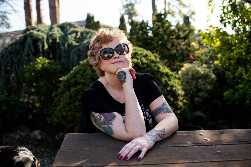 A woman with tattoos and dark glasses sits at a table in a garden.