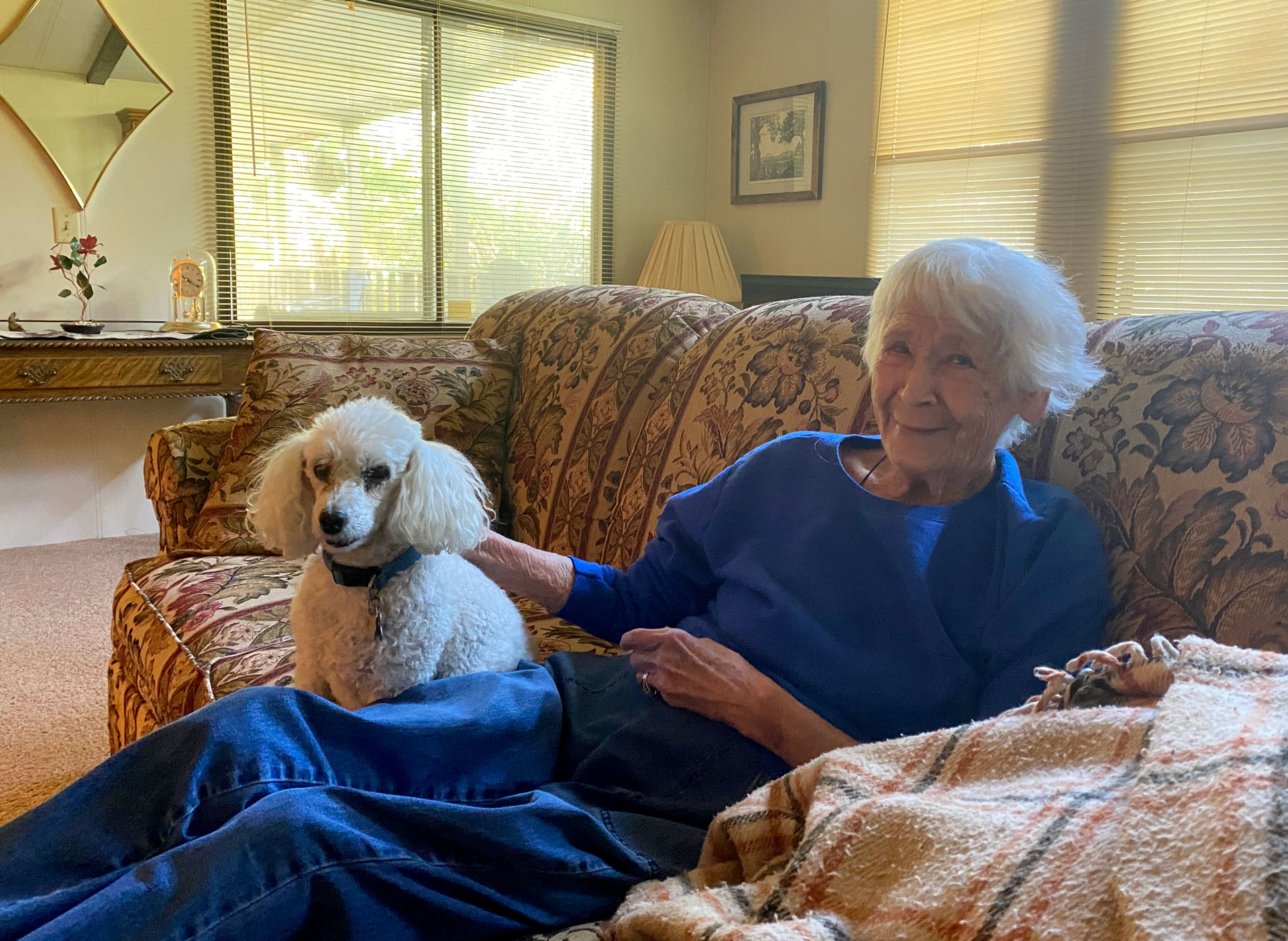 older woman wearing bright blug and smiling relaxes on her couch at home, petting a small white dog
