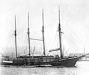 A black and white photo of a large boat with four masts, all sails furled. She rides low in the water, but her bow cuts through the waves.