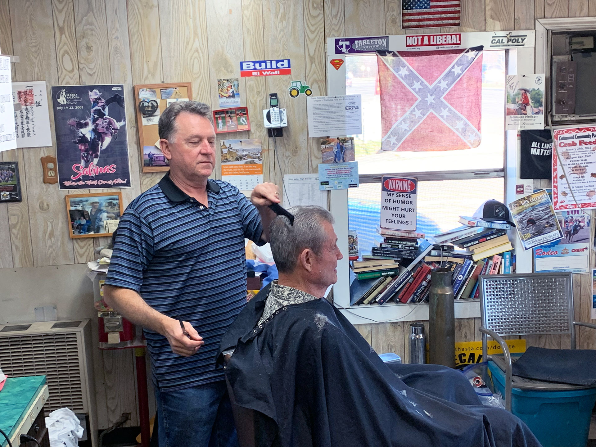 Barber combs man's hair with backdrop of confederate flag in the window, along with various bumper stickers including 'Build El Wall' and 'not a liberal' 