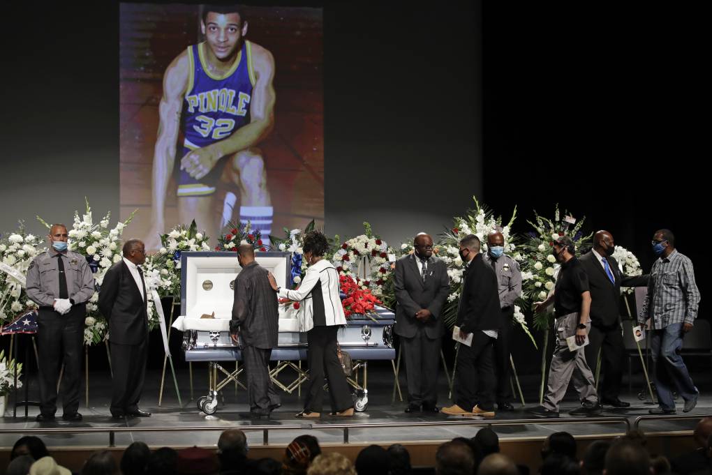 mourners file past an open casket funeral, a large photo of the deceased as a young man playing basketball appears in background