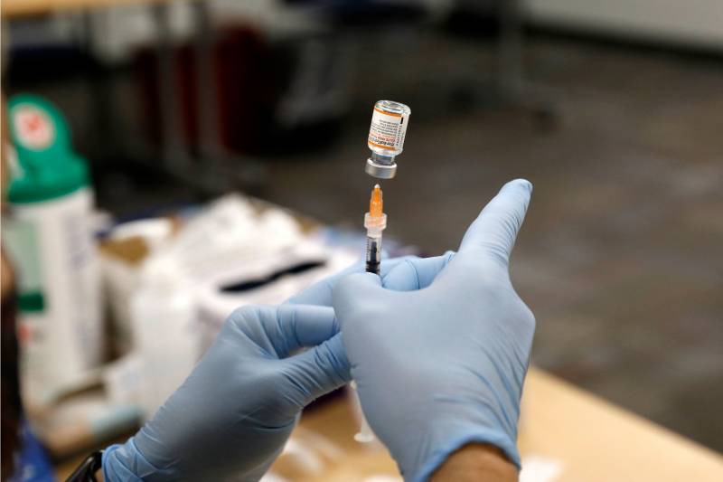 Two hands in light blue gloves hold a syringe inserted into an upside-down vial.