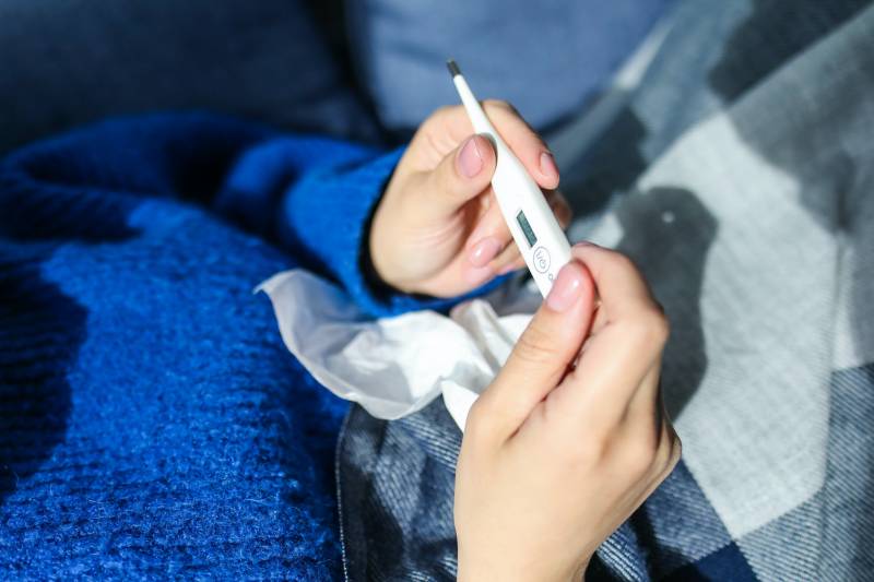 A person in a blue sweater holds a thermometer in their hands while laying under a knit blanket.