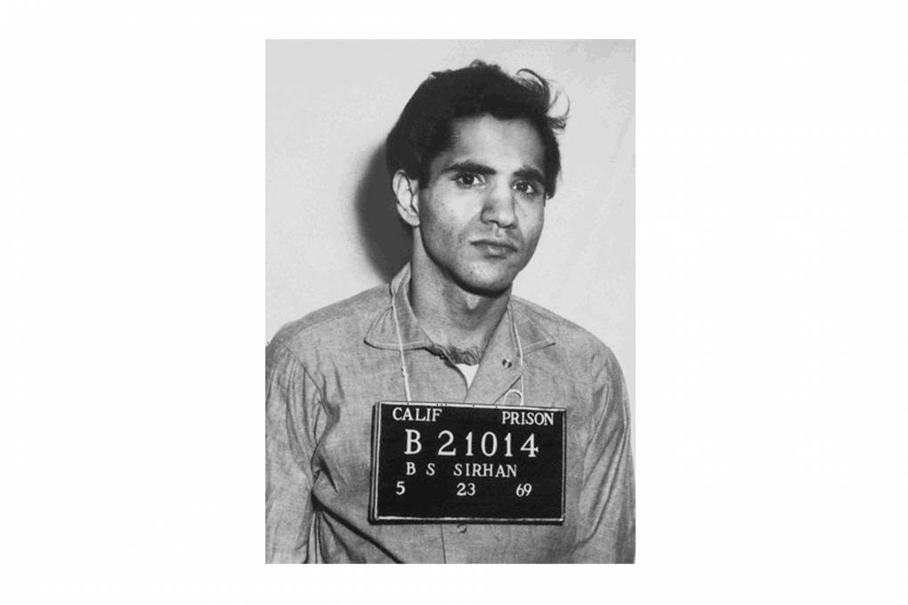 A black and white photo of a man wearing a collared shirt with a sign around his neck that reads "Calif Prison B21014 B S Sirhan"