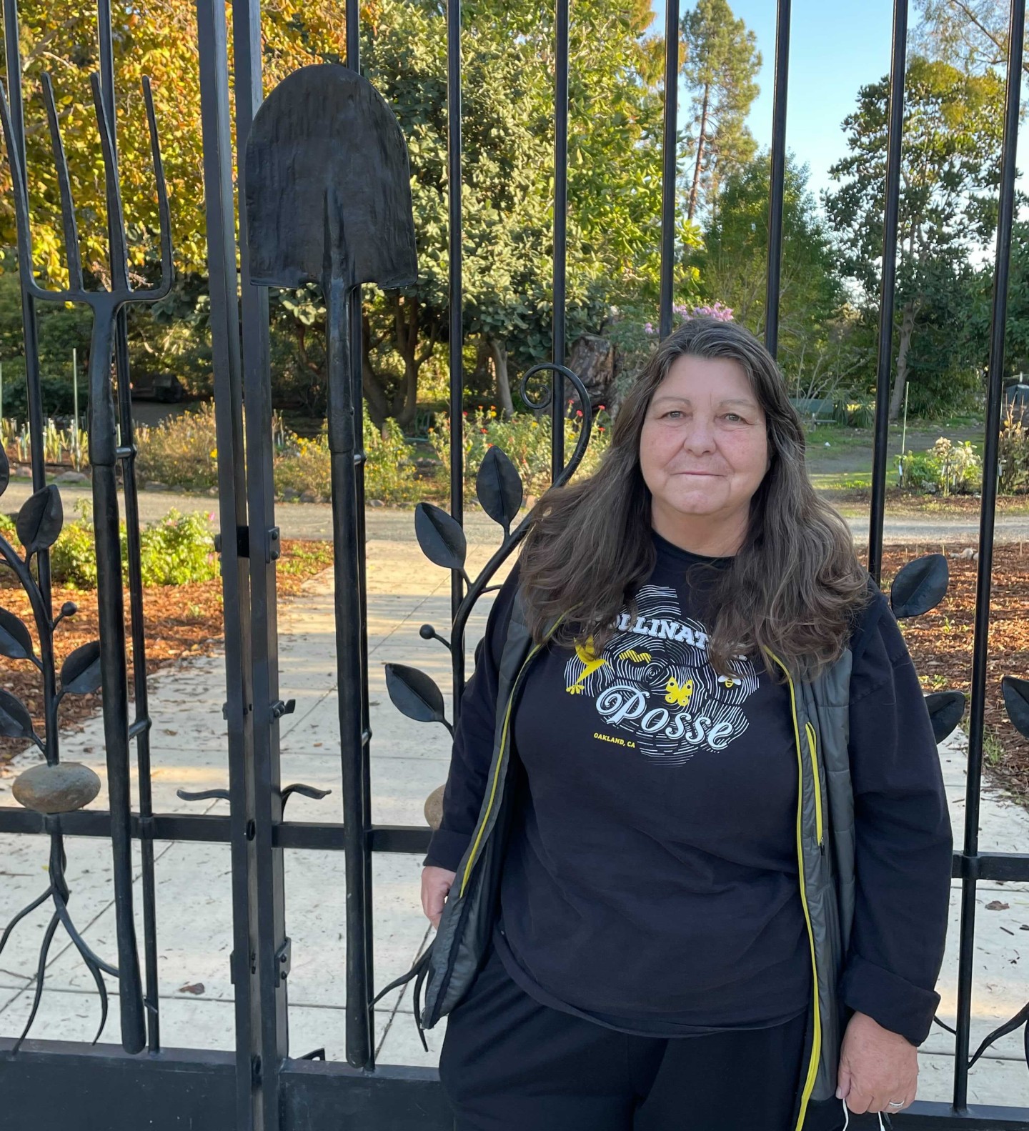A woman dressed in a shirt that says Polinator Posse on it stands in front of ornate iron gates leading to a garden.
