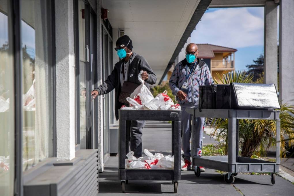 Two people push carts down a hotel hallway. The carts are full of food and bags and the people pushing the carts are wearing masks.