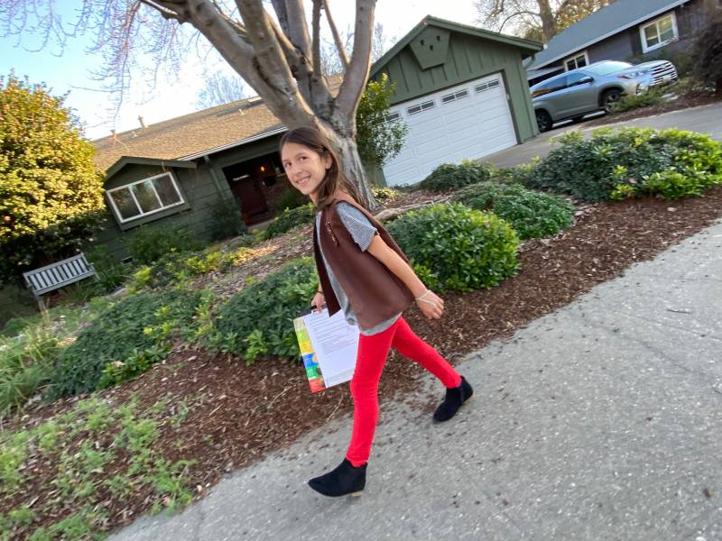 Girl scout wearing bright red pants and brown scouts vest smiles, carrying clipboard as she walks on a neighborhood street