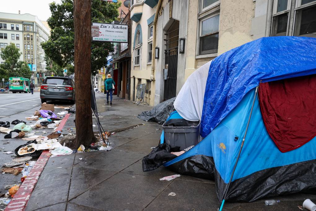 A blue tent has been set up along the wall of a building on a rainy sidewalk on a busy city street. The parking spaces nearest the tent are filled with trash – plastic bags, open takeout containers full of food, a paper cup – and additional trash is piled at the base of a tree next to the tent. In the background, a person in a green jacket walks away down the street.
