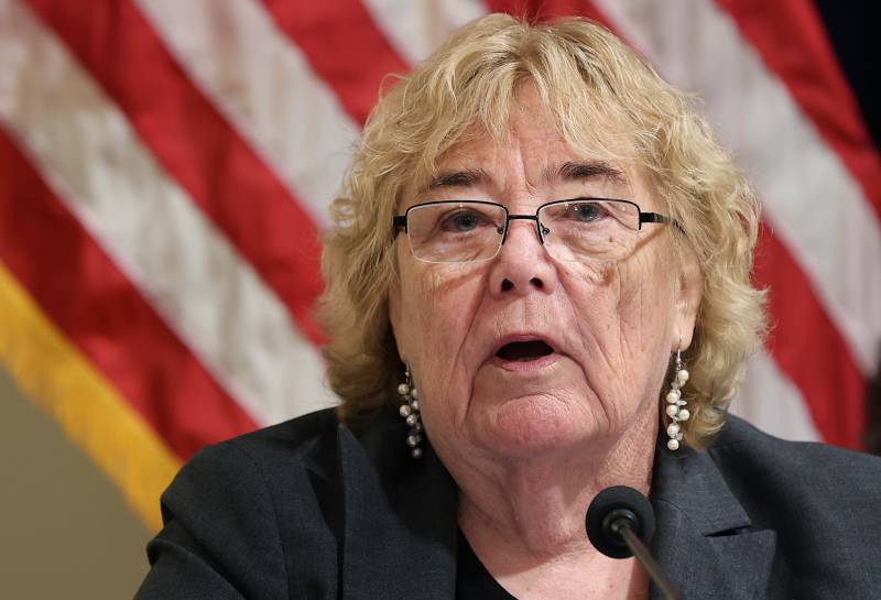 A close shot of Zoe Lofgren, a middle-aged white woman with shoulder-length blond hair and glasses, wearing large earrings and a dark blue blazer, speaks into a microphone in front of an American flag.