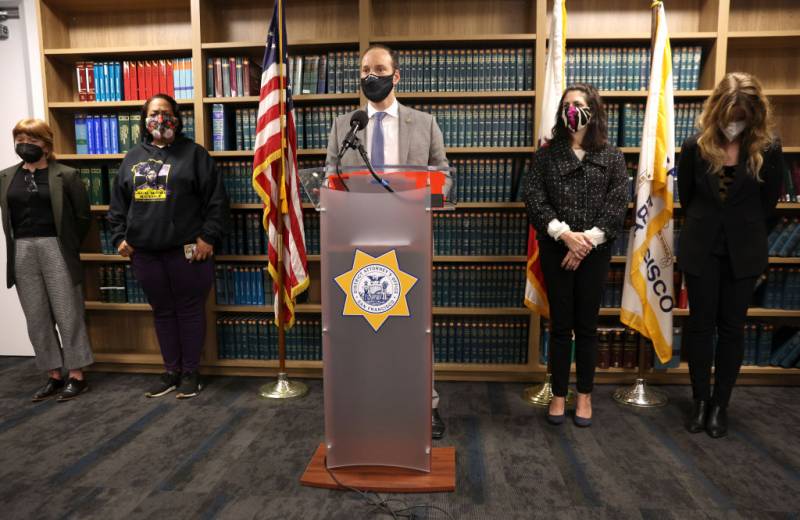 A man wearing a face mask stands behind a lectern, with two women on either side of him.