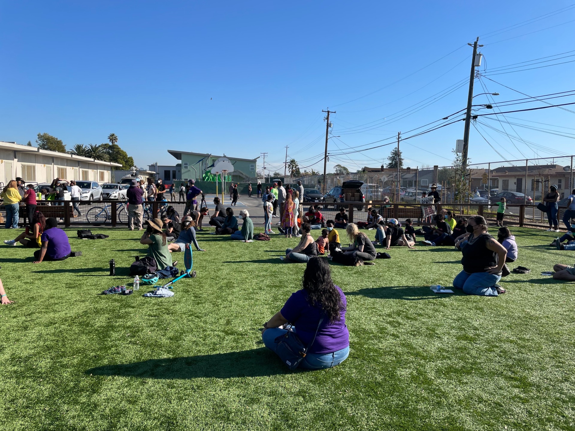 A few dozen people sit in the grass, many cross-legged, facing a speaker that is off-camera. A baskteball court and the street can be seen in the background.