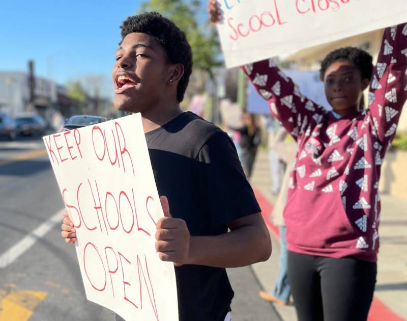 A young man, left, holds a picket sign reading "keep our schools open" while yelling to a passing car, a young woman holds another sign that is cut off from the photo, behind him, with blue skies behind them.