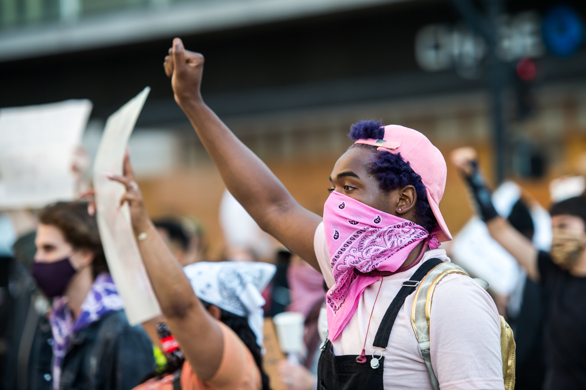 A young Black protester with pink bandana over their faces raises a fist along with other protesters