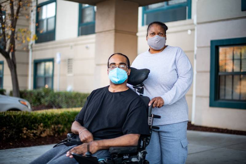 A mother and her son standing in front of a building. The woman is holding onto this wheelchair handles. They are both wearing masks.