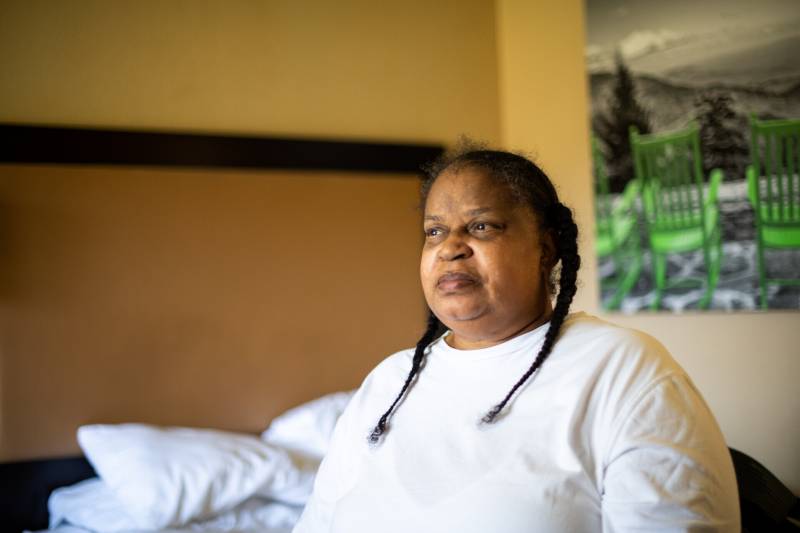 A woman stares off to the left while sitting on a bed. She is wearing a white shirt, and her hair is in braids.