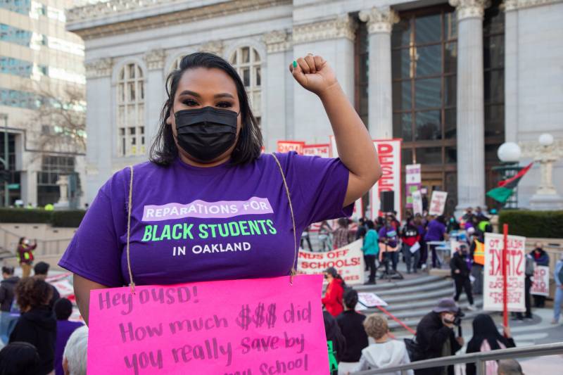 A woman in a purple shirt with the phrase 'Reparations for Black Students in Oakland' appears holding a pink protest sign with her fist up and Oakland City Hall in the background.