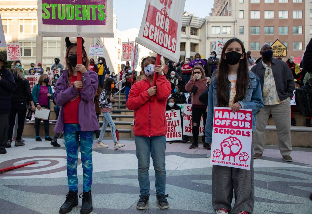 Three young people stand with their signs. One in a purple jacket, one in a red jacket and one person in a jean jacket with a sign saying "Hands off our schools."
