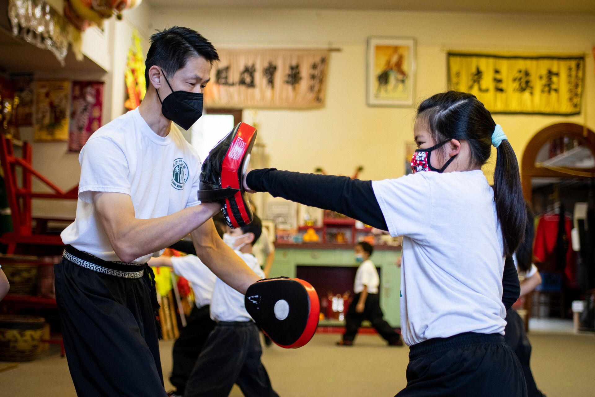 Kung fu instructor holds blocking pad while young girl student punches inside a kung fu studio