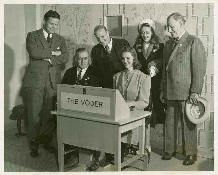 A woman plays a keyboard machine while 5 people look on.