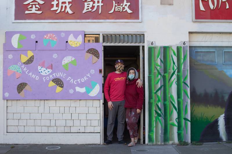 A woman and man stand in the frame of a doorway a red sign above and a sign marking the fortune cookie shop on the left