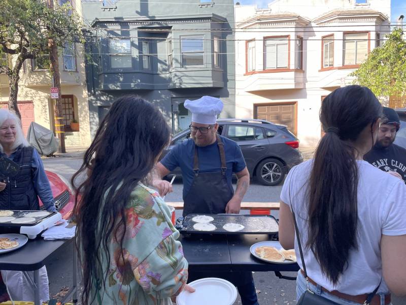 A man in a chef hat stands at a griddle on the sidewalk in San Francisco serving pancakes.