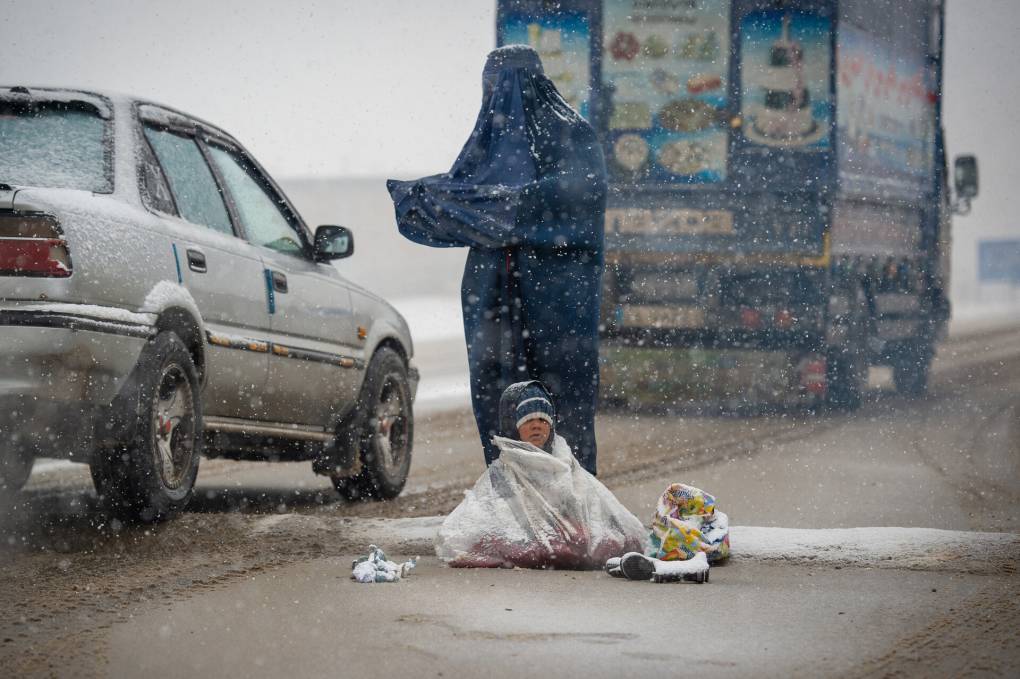 A woman begs for money from passing cars as a child sits beside her in the snow.