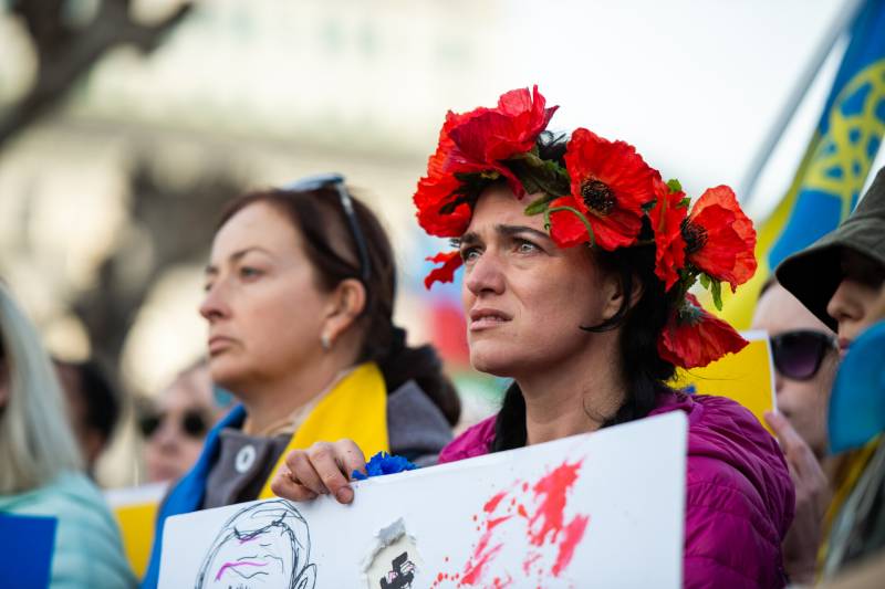 woman holding protest sign wearing crown of red flowers looks intently with teary eyes into the distance