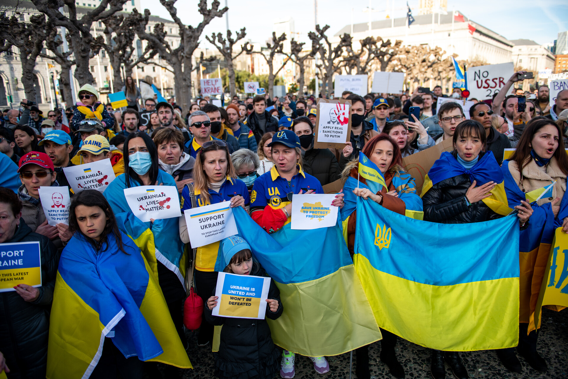 A group of people stand together holding blue and yellow Ukrainian flags and small signs in support of Ukraine.
