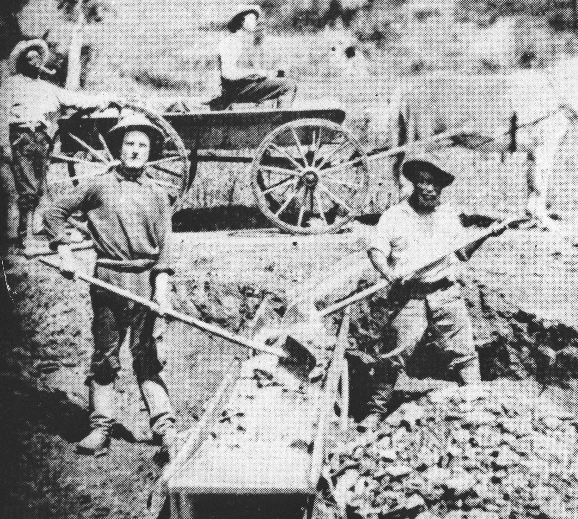Black and white image of men holding shovels standing next to a pile of rocks with a horse-drawn wagon in the background