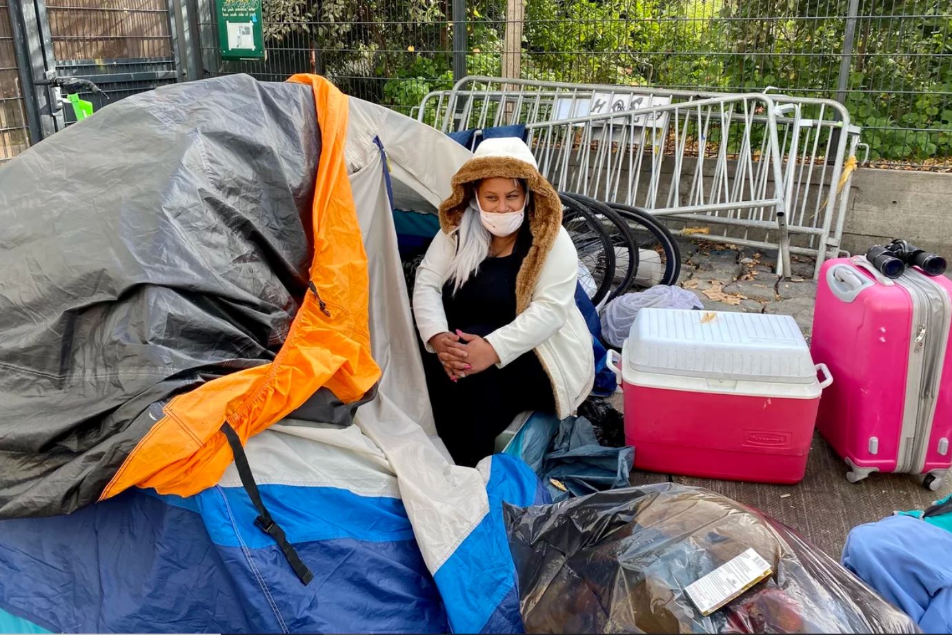 A person wearing a winter jacket and a face mask sits on the door of a camping tent that is set up on a sidewalk in San Francisco.