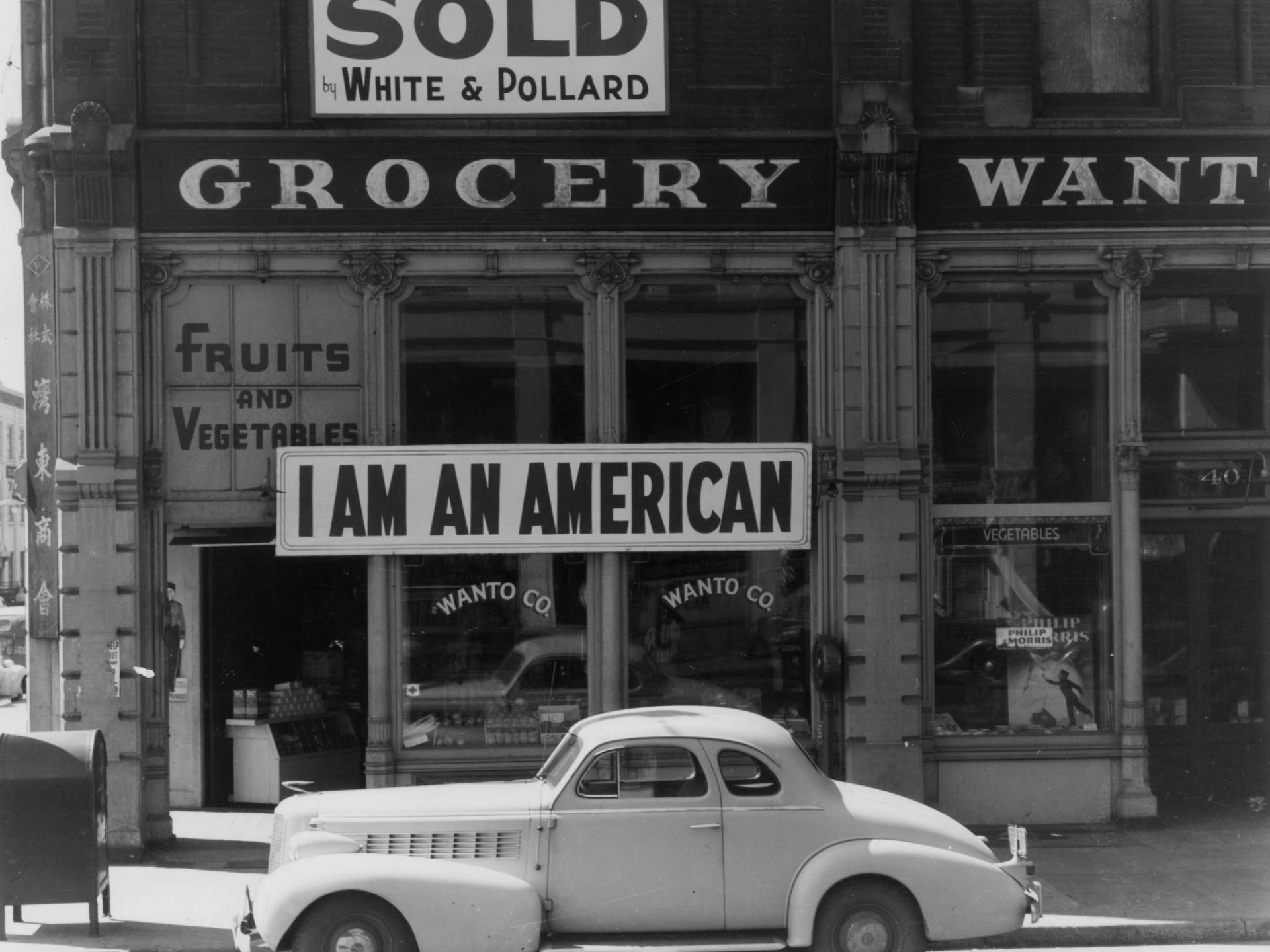 black and white photo of a car in front of a downtown Oakland storefront, with a large sign saying 'I AM AN AMERICAN' hung above the entrance