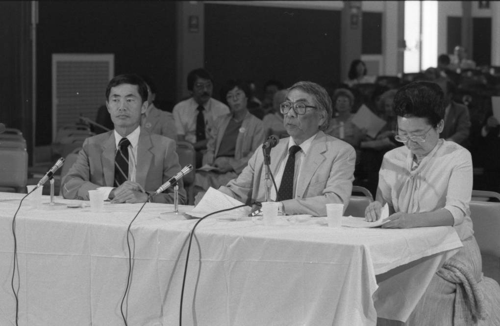 Black and white photo of two men and one woman sitting at a desk with microphones in front of them