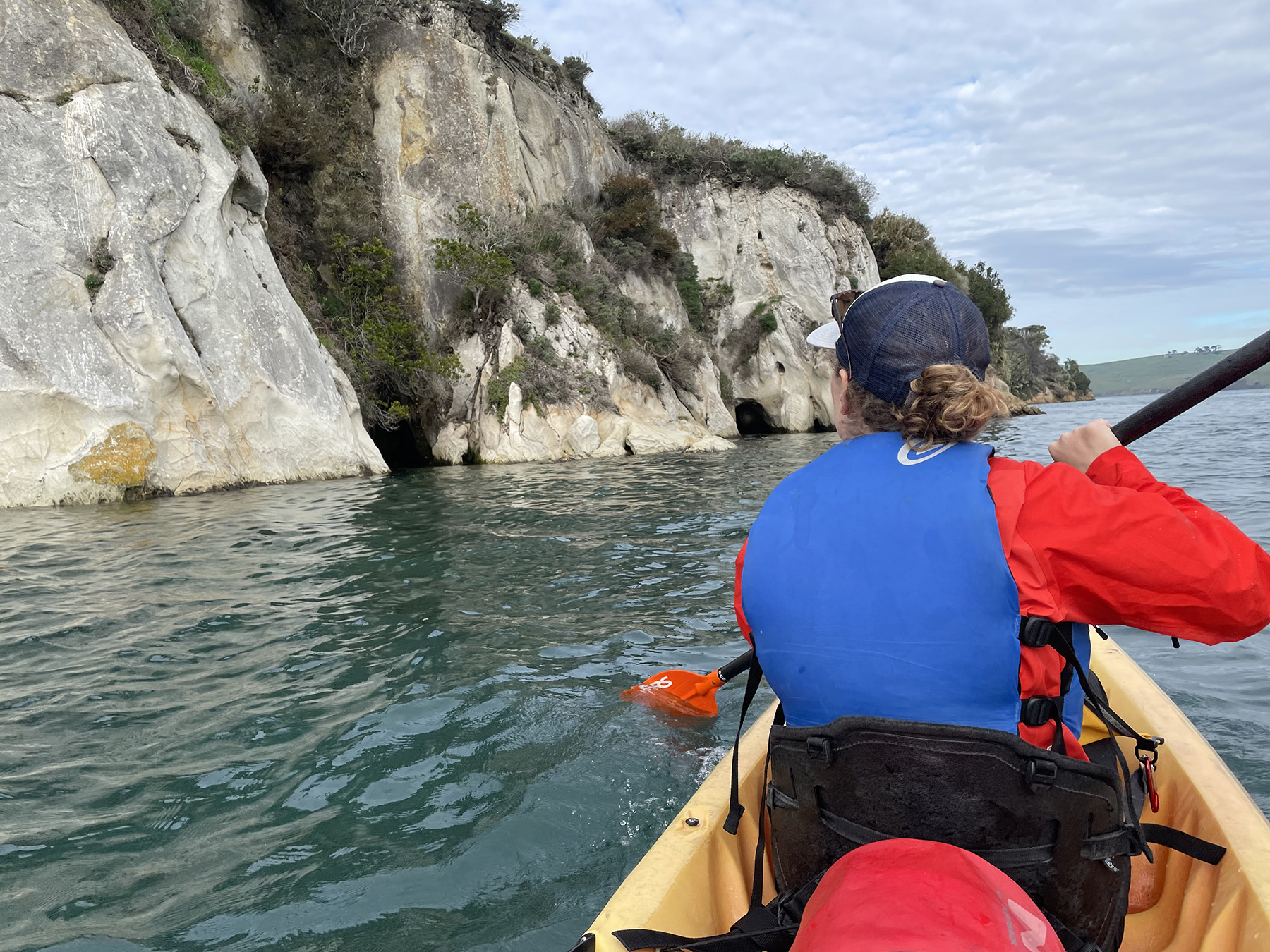 A woman in a red long sleeved shirt and blue life vest sits in the fron tof a kayak paddling towards a cliff in the distance.