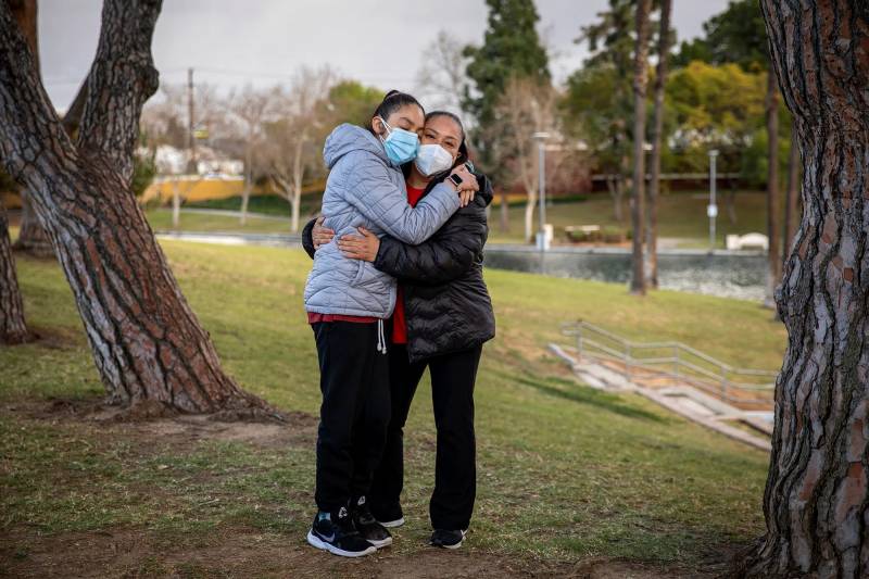 Two women wearing face masks hug each other in a park.