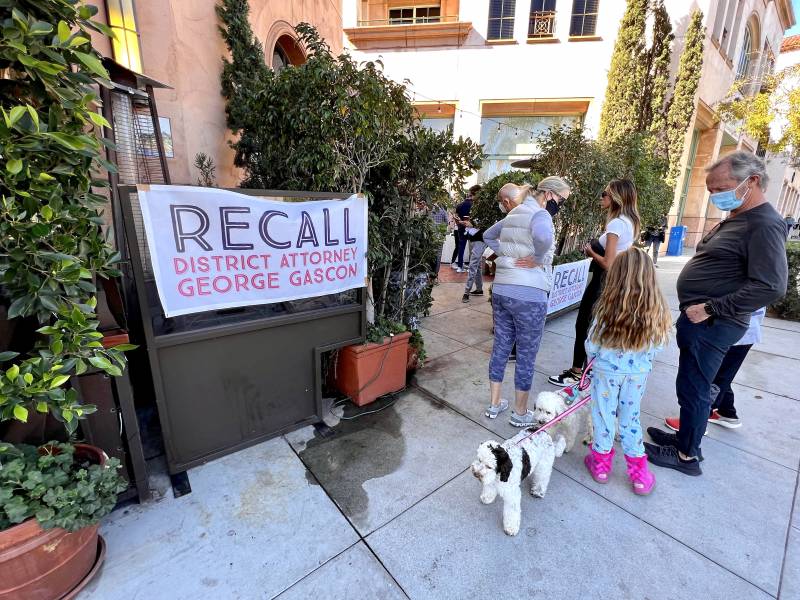 A 'Recall District Attorney George Gascón" banner hangs on a street, as several people - including a kid with a dog - stand nearby.