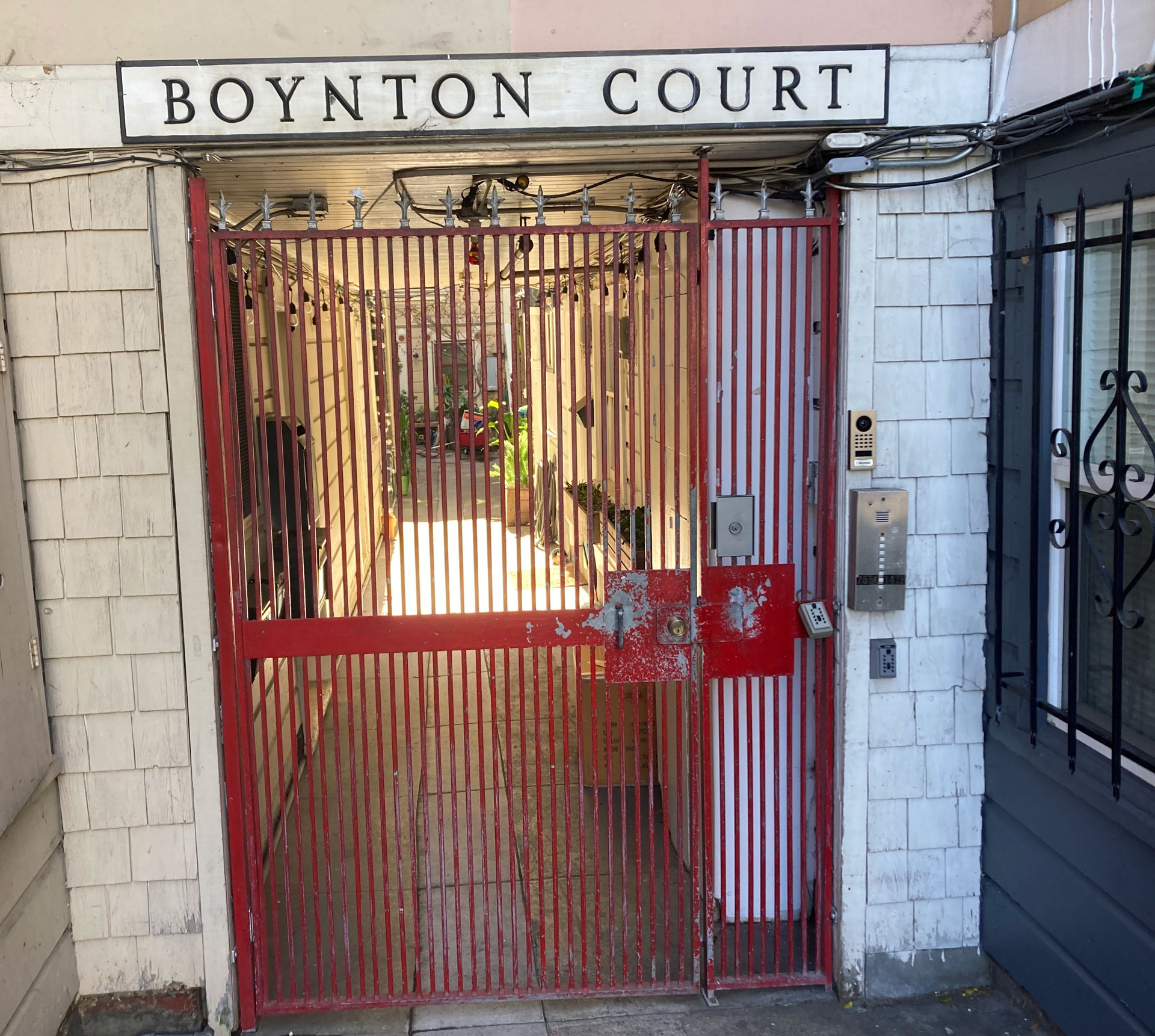 Through a red metal gate a passageway can be glimpsed with a sunny courtyard beyond. Above the gate a sign reads Boynton Court.