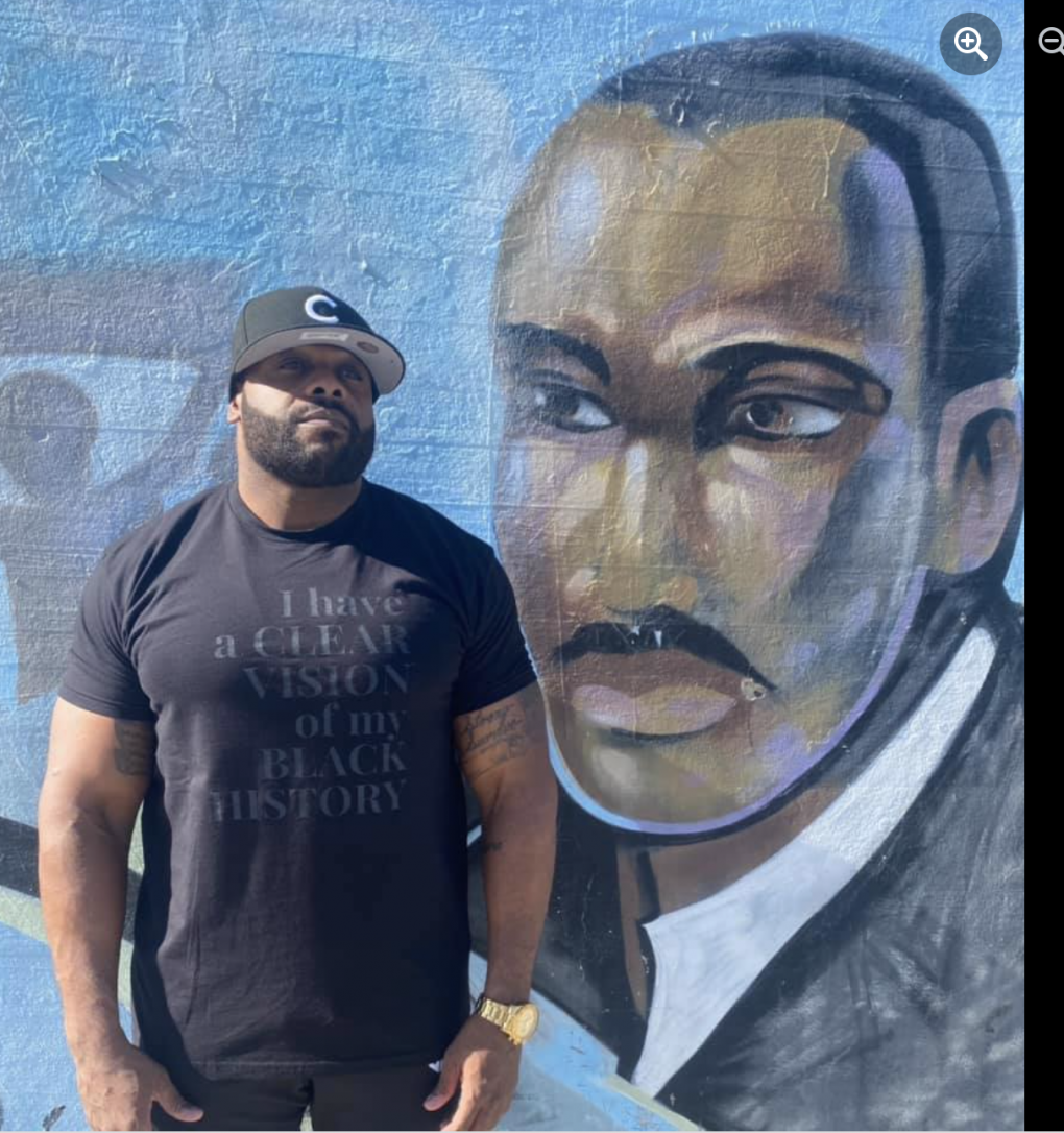 A man wearing a black baseball cap and black t shirt stands in front of a mural.