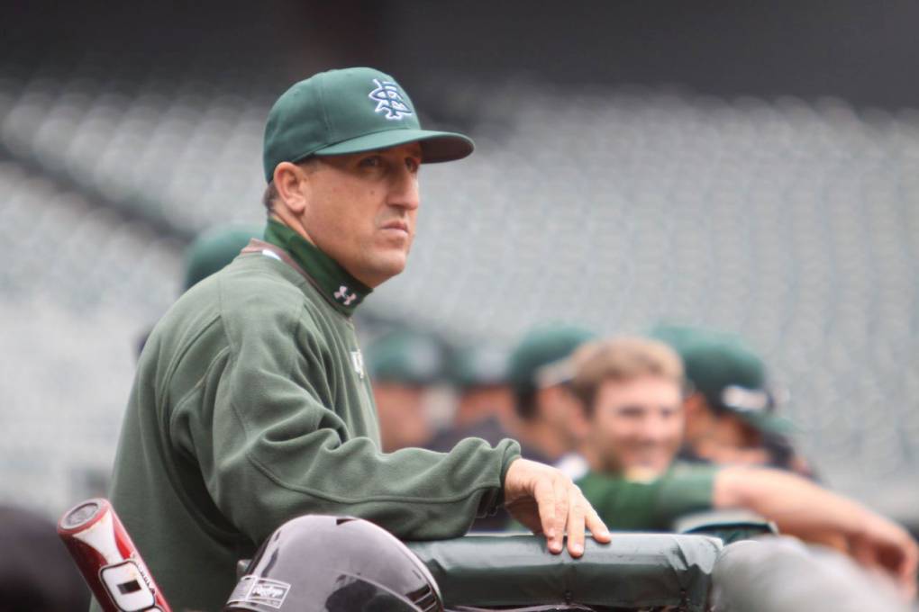 Profile photo of coach wearing green, standing in dugout, looking concerned