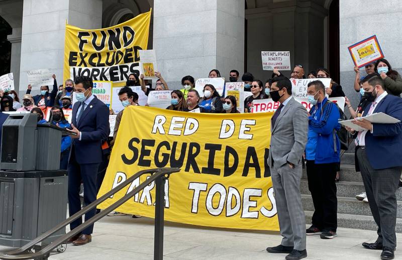 man in suit speaks at podium as ralliers hold bright yellow signs reading 'fund excluded workers'