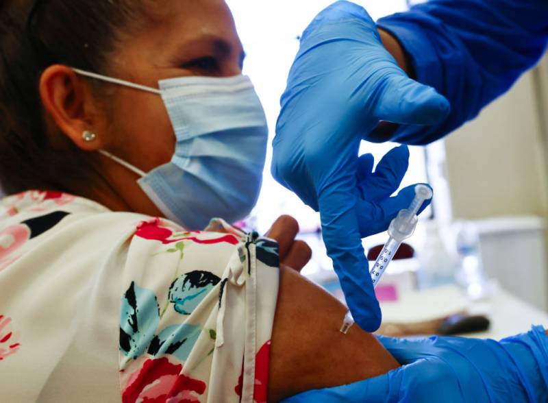A woman wearing a face mask receives an injection in her arm from a needle by a person wearing blue surgical gloves.