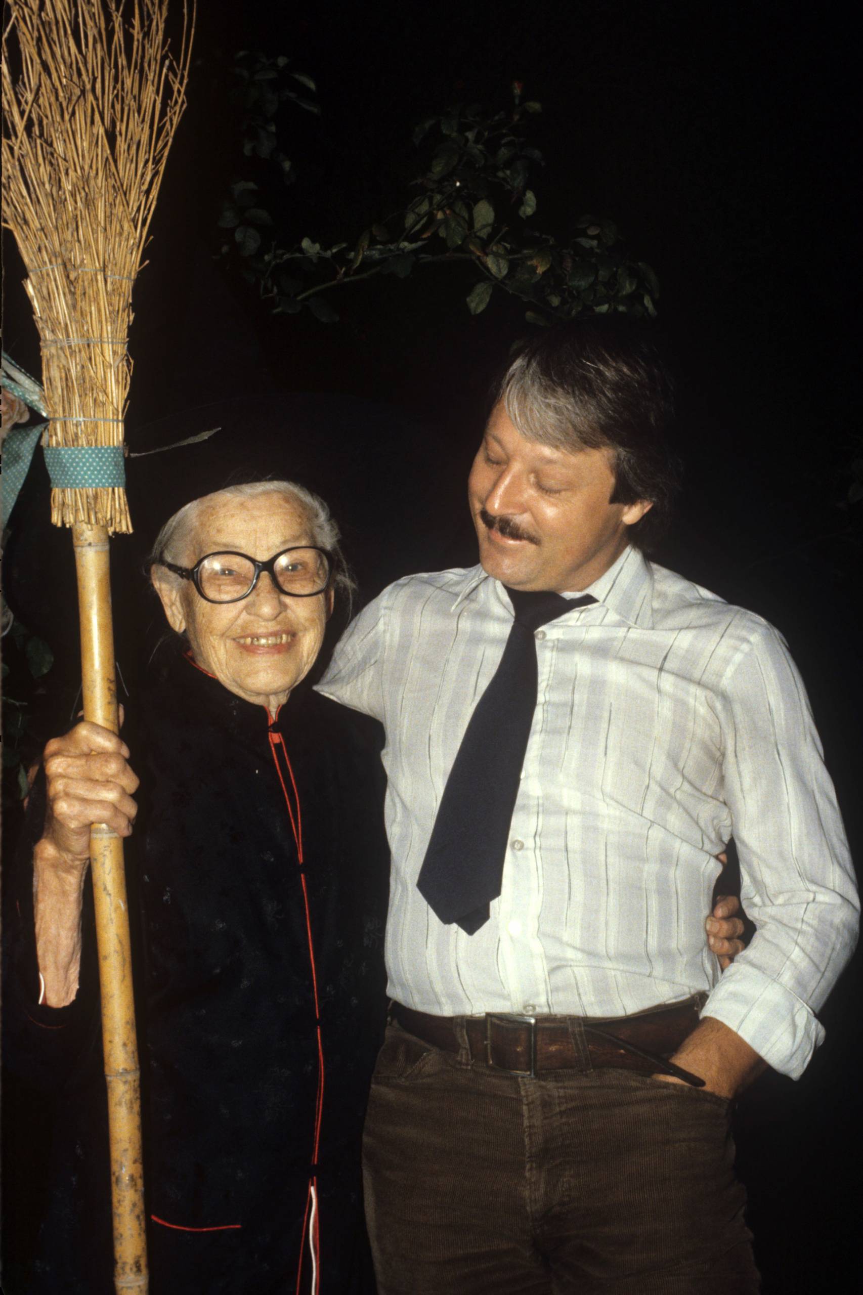 A posed photo of an older woman in a witch costume holding a broom. Next to her is a younger man with a mustache looks down fondly at the woman.