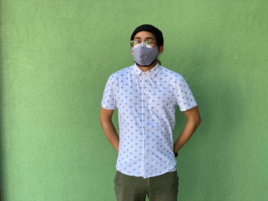 A school teacher stands against a green wall, wearing a mask, with his arms folded behind his back, posing for a portrait.