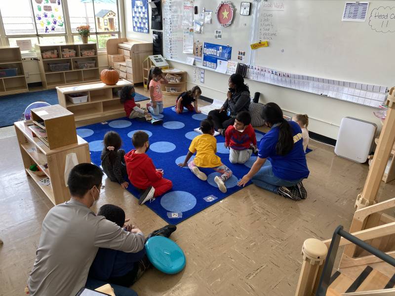 Children and adults sit on the floor in a classroom.