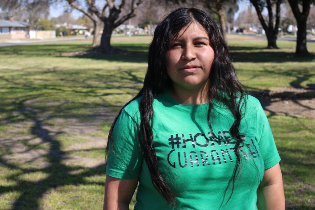 A woman stands in a park, wearing a green shirt that says, "#HomesGuarantee!""