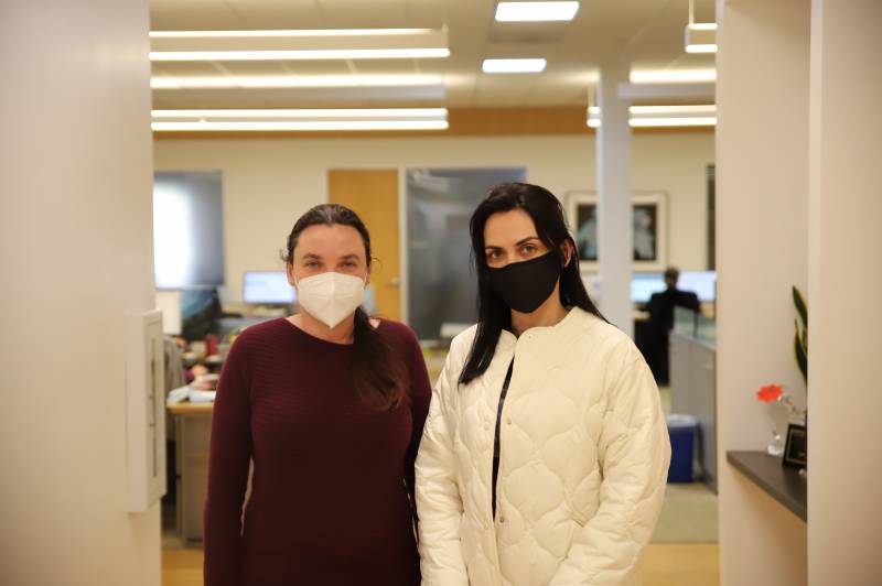 Two women stand together in a building. The woman on the right has a black face mask and a white jacket. The woman on the left has a white face mask and dark-colored shirt.